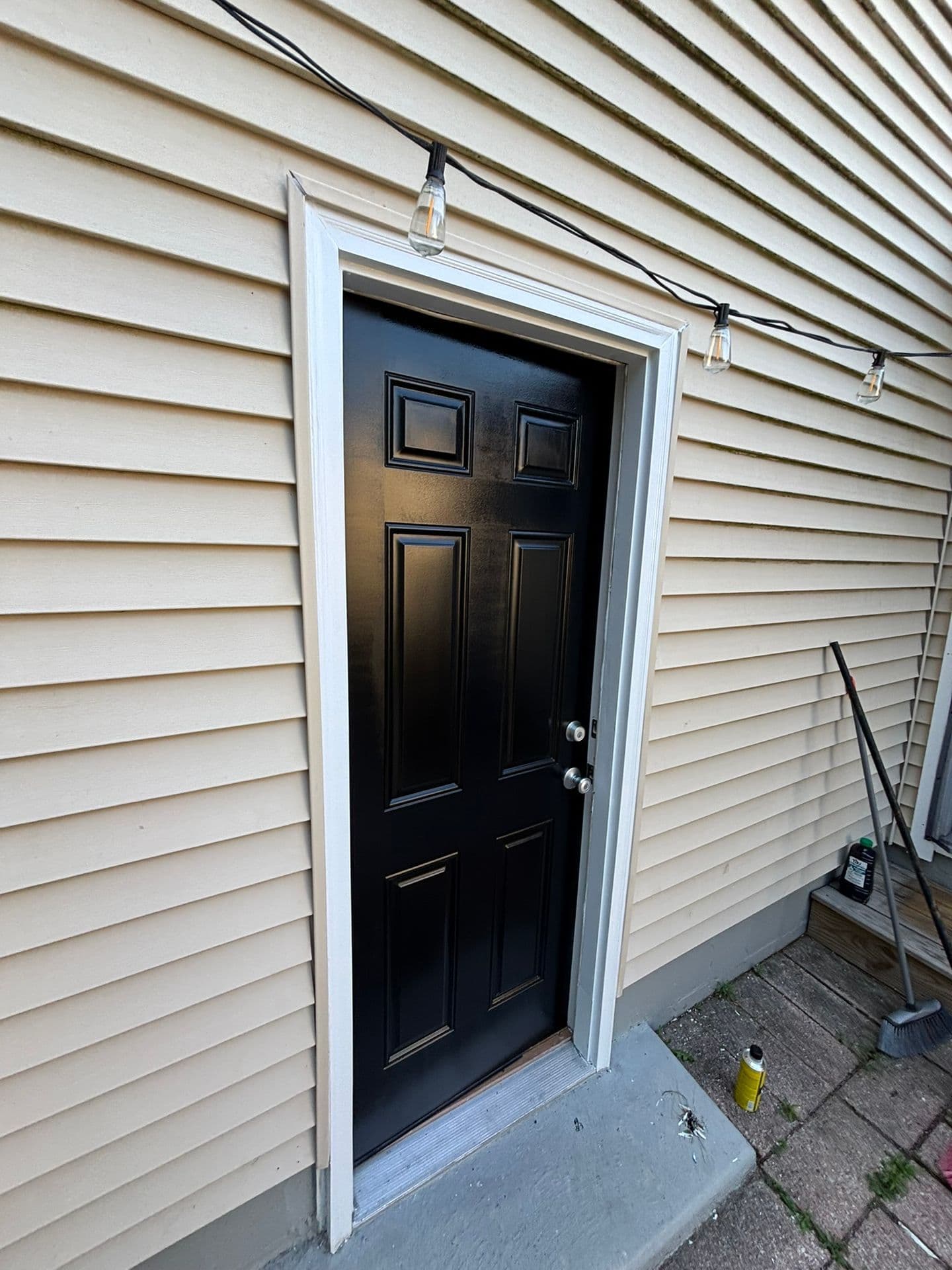 A black six-panel exterior door with white trim set against beige horizontal siding, decorated with string lights above.